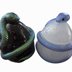 May include: Two decorative, snake-themed containers. One is black with a green and blue iridescent snake wrapped around it. The other is clear with a blue snake. Both have a rounded shape and are likely used for storage.
