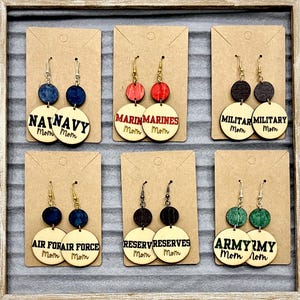 May include: Wooden dangle earrings with various military branch themes. Each pair features a circular wooden base with text like "NAVY Mom" or "MARINES Mom", and a smaller wooden circle above. The earrings are displayed on brown cards.