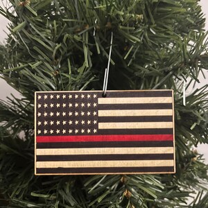 May include: Wooden ornament shaped like the American flag with a thin red stripe across the middle. The ornament is hanging on a Christmas tree.