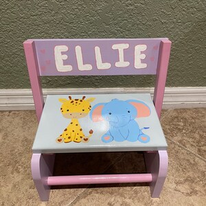 Puede incluir: Un taburete de madera rosa con una parte superior gris que presenta una jirafa y un elefante pintados. El taburete tiene un pasamanos rosa y el nombre "ELLIE" está pintado en la parte posterior.