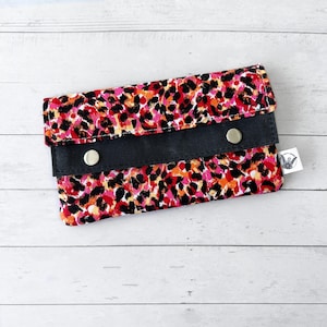 May include: A rectangular fabric pouch featuring a vibrant leopard print pattern in shades of pink, orange, and black. The pouch has a horizontal black fabric band across the front secured by two silver-toned metal snap buttons and a small white tag.