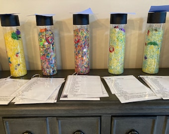 Puede incluir: Cinco tarros de vidrio transparente con tapas negras y gorros de graduación azules en la parte superior, llenos de arroz de colores y pequeños objetos. Cada tarro tiene una hoja de papel blanca con el texto "I Spy Jar" y una lista de objetos para encontrar.
