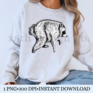 Peut inclure: Un sweat-shirt gris clair &agrave; col rond orn&eacute; d'une illustration au trait noir et blanc repr&eacute;sentant un raton laveur de profil. Le raton laveur est repr&eacute;sent&eacute; en position de marche avec une texture de fourrure d&eacute;taill&eacute;e. Le texte 28th Design Co. est visible.