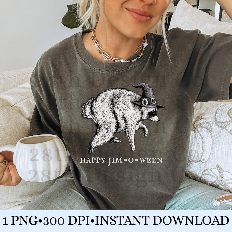 May include: A charcoal gray short-sleeve t-shirt featuring a black and white illustration of a raccoon wearing a pointed witch hat. Below the raccoon, the text reads HAPPY JIM-O-WEEN in white capital letters. The shirt has a crew neckline.