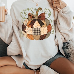 May include: A cream-coloured long-sleeved shirt with a patchwork pumpkin design. The pumpkin is made up of various fabric patterns in shades of brown, orange, and beige, with a dark brown bow and green vine accents. A fall-themed graphic tee.