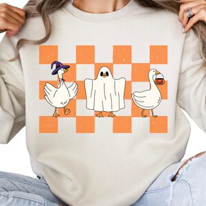 May include: Cream-colored sweatshirt featuring a Halloween-themed design. The graphic includes three cartoon geese: one wearing a witch hat, one dressed as a ghost, and one carrying a jack-o'-lantern bucket. The background is a checkered pattern in orange and white.