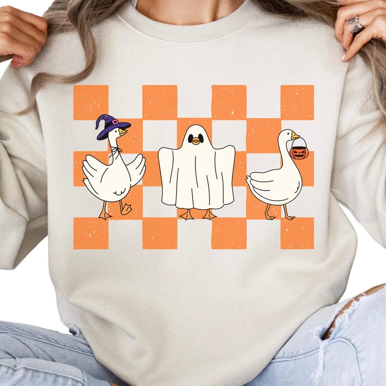 May include: Cream-colored sweatshirt featuring a Halloween-themed design. The graphic includes three cartoon geese: one wearing a witch hat, one dressed as a ghost, and one carrying a jack-o'-lantern bucket. The background is a checkered pattern in orange and white.