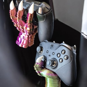 May include: A pair of iridescent, claw-shaped controller holders, one holding a black Xbox controller. The holders are in shades of purple, green, and gold, with white claw tips. The text "3Deality Designs" is visible.