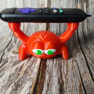 Op de afbeelding: Een rode 3D-geprinte krab met groene ogen die een zwarte afstandsbediening vasthoudt.