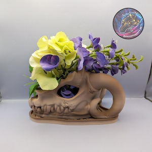 May include: A tan-colored, 3D-printed dragon skull planter filled with yellow and purple artificial flowers. The planter has a detailed design with horns and eye sockets. A circular sticker with a horse head logo is visible in the background.