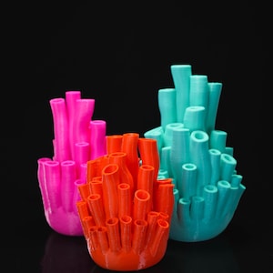 May include: Three 3D printed decorative sculptures shaped like coral reefs, featuring a bright pink, a vibrant orange, and a teal blue piece. Each sculpture consists of multiple vertical, hollow, tube-like structures with textured surfaces.
