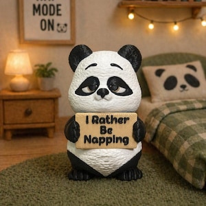 Pode incluir: Uma estatueta de panda em preto e branco com superfície texturizada, segurando uma placa retangular bege com a inscrição "I Rather Be Napping" em letras pretas. O panda tem olhos sonolentos e está em pé com orelhas e membros pretos.