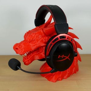 Puede incluir: Un soporte para auriculares en forma de dragón rojo con un auricular para juegos negro y rojo apoyado en él. El auricular tiene un micrófono.