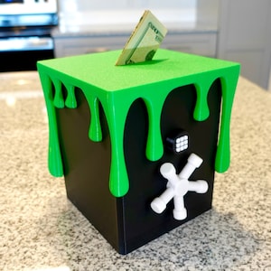 Può includere: Un salvadanaio quadrato nero con un design a goccia di melma verde sulla parte superiore e sui lati. Il salvadanaio ha una serratura a combinazione bianca e una maniglia bianca a forma di stella. Una banconota da venti dollari è inserita nella fessura.