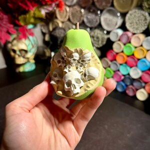 Può includere: Una scultura a forma di pera verde con un gruppo di teschi bianchi che emergono dal centro. La pera ha una superficie strutturata e un piccolo gambo marrone. La scultura è tenuta in mano, con uno sfondo sfocato di oggetti colorati.