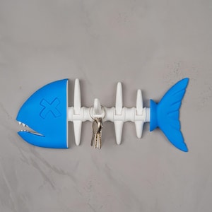 May include: A blue and white fish-shaped key holder with a cartoonish design. The key holder has a blue head and tail, with a white spine and key hooks. A set of keys hangs from the hooks. The key holder is mounted on a gray wall.