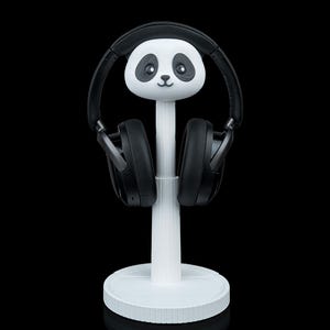 May include: A white panda-shaped headphone stand with black headphones. The stand has a round base and a vertical support topped with a panda head. The headphones are black and rest on the panda's ears.