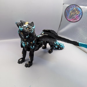 May include: A black fantasy creature figurine with wings, adorned with teal and gold accents. The wolf-like figure has a crescent moon on its forehead and intricate patterns. The wings are black with teal details. A circular logo is in the background.