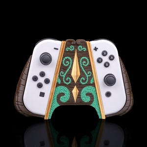 Może przedstawiać: Niestandardowy kontroler do gier wideo z białymi bokami i brązowo-zielonym panelem środkowym. Środkowy panel ma fakturę przypominającą drewno z zielonymi wirami i złotymi akcentami. Kontroler ma czarne przyciski i joysticki.