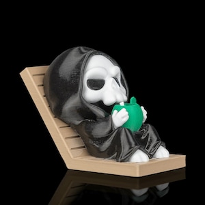 Peut inclure: Une petite figurine représentant un personnage au crâne blanc vêtu d'une robe à capuche noire, assis sur une chaise longue à lattes beige. Le personnage tient dans ses mains un récipient à boisson vert vif, rond et texturé, avec une paille blanche.