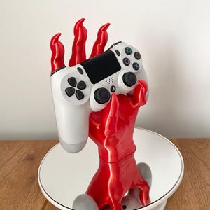 May include: A white PlayStation 4 controller rests in the grasp of a glossy red hand-shaped stand. The stand is on a white mirrored surface, reflecting the controller and the red hand. The controller has black buttons and a black touchpad.