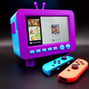 Puede incluir: Una consola retro en forma de televisor de color morado y azul con una pantalla que muestra un menú de Nintendo Switch. Dos mandos de Nintendo Switch azules y naranjas están en primer plano.