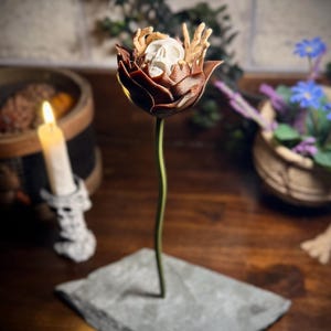 May include: A brown ceramic flower with a white skull and skeletal hands inside. The flower is on a grey stone base.