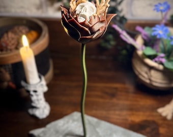 May include: A brown ceramic flower with a white skull and skeletal hands inside. The flower is on a gray stone base.