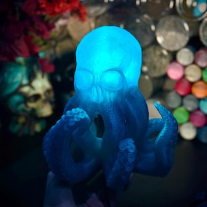 Puede incluir: Lámpara azul con forma de calavera y tentáculos de pulpo. La lámpara emite una luz azul brillante, resaltando los detalles del cráneo y los tentáculos. Un artículo decorativo único.