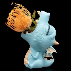 May include: A light blue dragon-shaped vase with a large orange flower and green foliage emerging from its mouth. The dragon has tan accents on its claws, wings, and belly. The background is black.