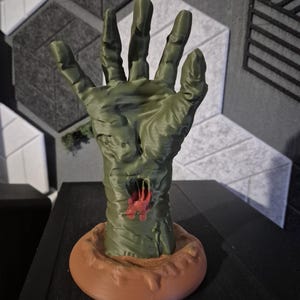 Op de afbeelding: Een groene, 3D-geprinte zombiehand met een gescheurde wond, rustend op een bruine cirkelvormige basis. De hand is gedetailleerd met een getextureerde huid en open vingers. De basis staat op een donker oppervlak.