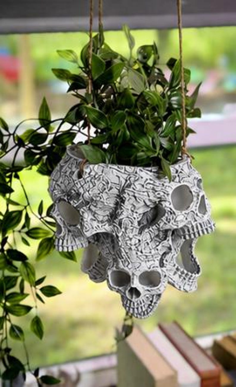 May include: A hanging planter featuring a cluster of white and grey skulls with intricate, dark, etched detailing. The planter is suspended by thin brown cords and contains a lush green plant with pointed leaves that spills over the edges.