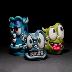 Op de afbeelding: Drie 3D-geprinte monster-vormige houders. Links een hoge blauwe houder met paarse stippen en meerdere ogen. In het midden een kortere, grijsblauwe creatuur met grote ogen en tanden. Rechts een groene houder met grote ogen en een rode tong.