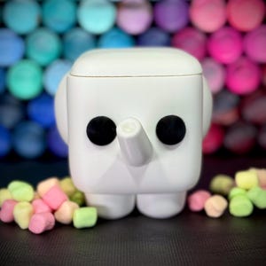 May include: A white, square-shaped elephant figurine with black eyes and a trunk. The figurine has rounded ears and feet. Colorful marshmallows are scattered in front of the elephant against a blurred background of colorful spheres.