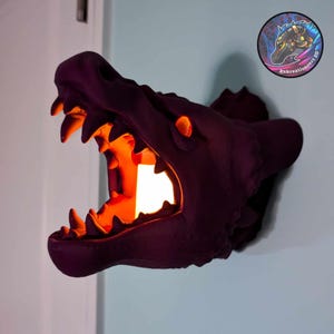 Könnte beinhalten: Ein lilafarbener 3D-gedruckter Drachenkopf-Wandhalter mit offenem Maul und leuchtendem orangefarbenem Licht im Inneren. Der Drachenkopf ist an einer weißen Wand montiert.