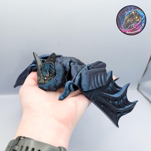May include: A dark blue bat figurine with detailed wings and a textured body. The bat is lying down, with its wings spread. The figurine has a realistic appearance and is likely 3D-printed. A sticker with a logo is visible in the background.