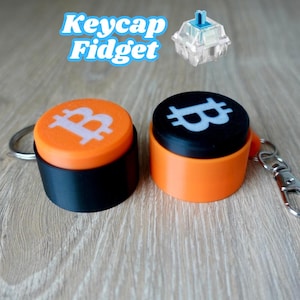 May include: Two orange and black keycaps with a Bitcoin symbol on the top. The keycaps are attached to a silver keychain. The text "Keycap Fidget" is written in blue letters above the keycaps.