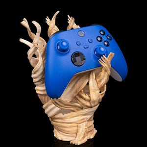 Peut inclure: Une manette Xbox bleue est présentée, tenue par une sculpture beige texturée. La manette a des boutons noirs et une croix directionnelle noire. La sculpture a un aspect rugueux et organique, sur fond sombre.