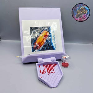 Könnte beinhalten: Ein lavendelfarbenes Diamantmalerei-Set mit einem teilweise fertigen Fischbild. Das Set enthält eine Schale mit roten Edelsteinen, einen Stift und einen kleinen Behälter mit roten Edelsteinen. Das Bild befindet sich in einer transparenten Plastikhülle.
