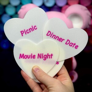 以下が含まれることがあります： ピンク色の文字で「Picnic」、「Dinner Date」、「Movie Night」と書かれた白いハート型のカード。手で持ち、背景にはカラフルなオーブとピンク色の形がぼやけています。