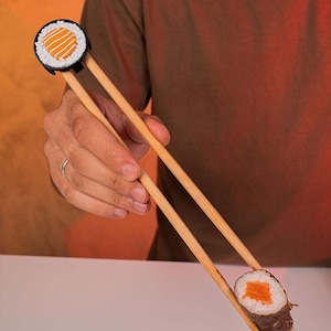 Könnte beinhalten: Essstäbchen aus Holz, die ein Stück Sushi halten. Die Essstäbchen haben oben einen schwarzen Sushi-förmigen Halter. Die Sushi-Rolle hat eine orangefarbene und weiße Füllung. Der Hintergrund ist ein Farbverlauf aus Orange und Braun.