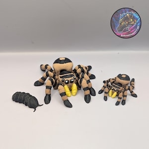 Puede incluir: Tres arañas de juguete impresas en 3D y un pequeño gusano negro segmentado. Las arañas son de rayas beige y negras con colmillos amarillos. La araña más grande está en el centro, con una más pequeña a la derecha. El gusano está a la izquierda.