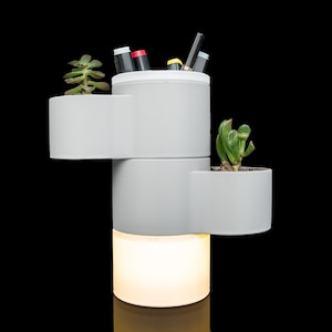 May include: A multi-tiered, light gray cylindrical organizer featuring three distinct sections for storage and planting. The top section holds pens, while the middle and upper side sections contain small green succulents. The base emits a warm white glow.