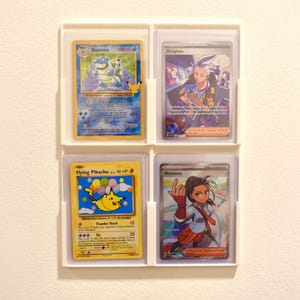 Op de afbeelding: Vier Pokémon-verzamelkaarten in beschermende hoesjes. De kaarten tonen personages als Blastoise, Flying Pikachu, Drayton en Nemona. Ze zitten in een witte display-frame, die de kleurrijke illustraties en tekst benadrukt.