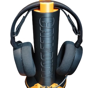 Könnte beinhalten: Ein schwarzer Gaming-Headset-Ständer mit orangefarbener Basis. Der Ständer trägt die Aufschrift "Call of Duty" an der Seite. Das Headset ist schwarz mit einem schwarzen Mikrofon.