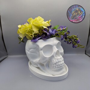 May include: A white skull-shaped planter with a matching saucer, filled with yellow and purple flowers. The planter has detailed facial features and is made of a smooth material. A circular logo with a horse head is in the background.