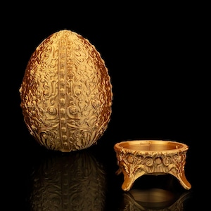 May include: A gold egg-shaped decorative box with intricate floral patterns and a matching gold stand with three legs. The box and stand are both made of metal and have a shiny finish.