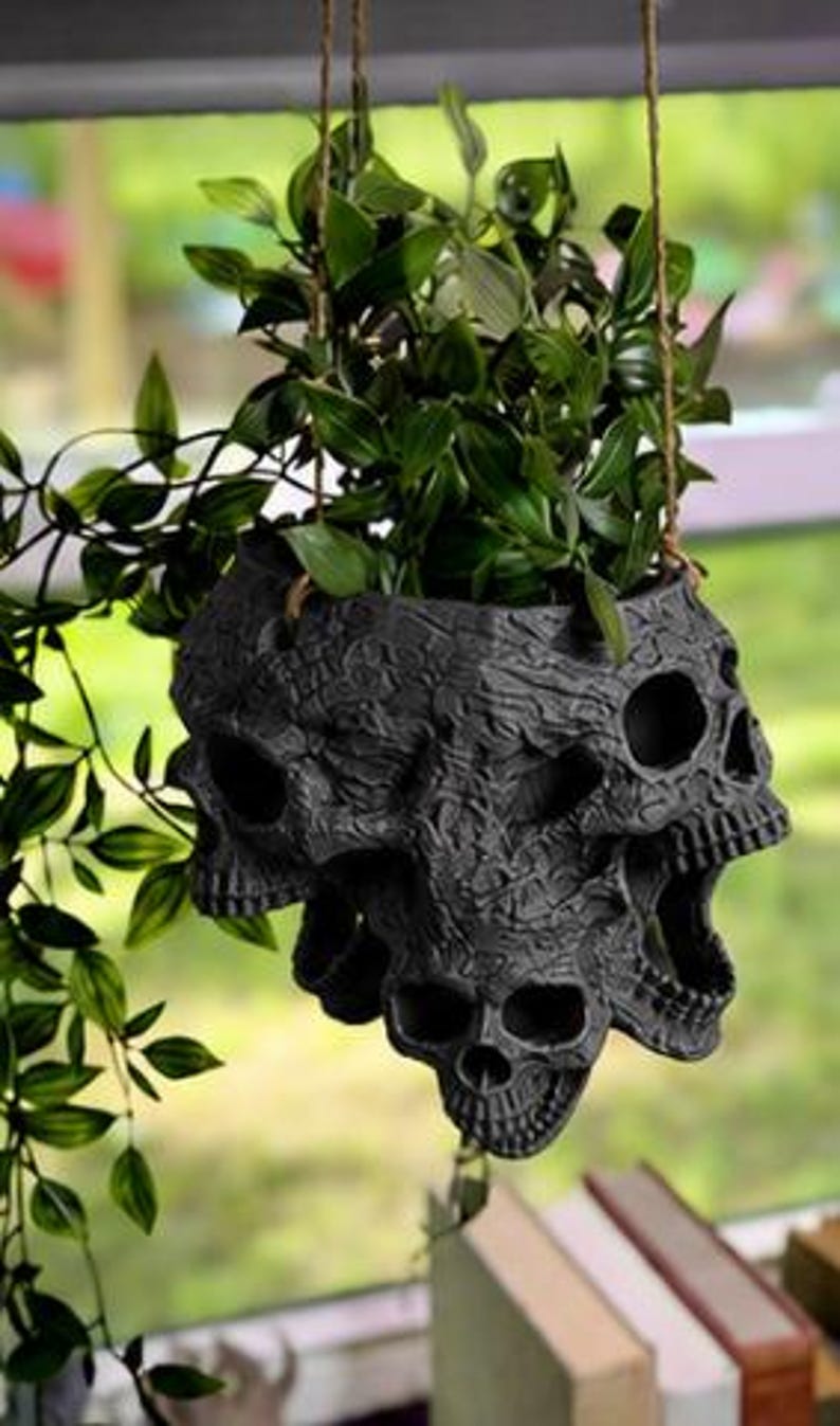 May include: A matte black hanging planter featuring a cluster of sculpted skulls with intricate textured details. The planter is suspended by thin brown twine and contains a lush green trailing plant with pointed leaves extending outward.
