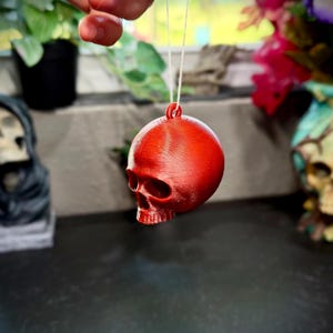 Könnte beinhalten: Ein kleiner, runder, roter Schädel-Ornament hängt an einem dünnen weißen Faden. Der Schädel hat detaillierte Gesichtszüge, einschließlich Augenhöhlen, einer Nasenhöhle und einem Kiefer. Das Ornament ist wahrscheinlich für Halloween oder Gothic-Dekoration.