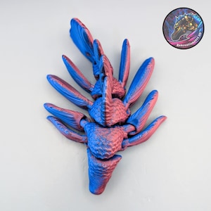 May include: A 3D-printed sculpture in shades of blue and red, resembling a stylized organic form. The object has multiple curved, wing-like appendages radiating from a central core. A circular logo with a horse head is in the upper right corner.